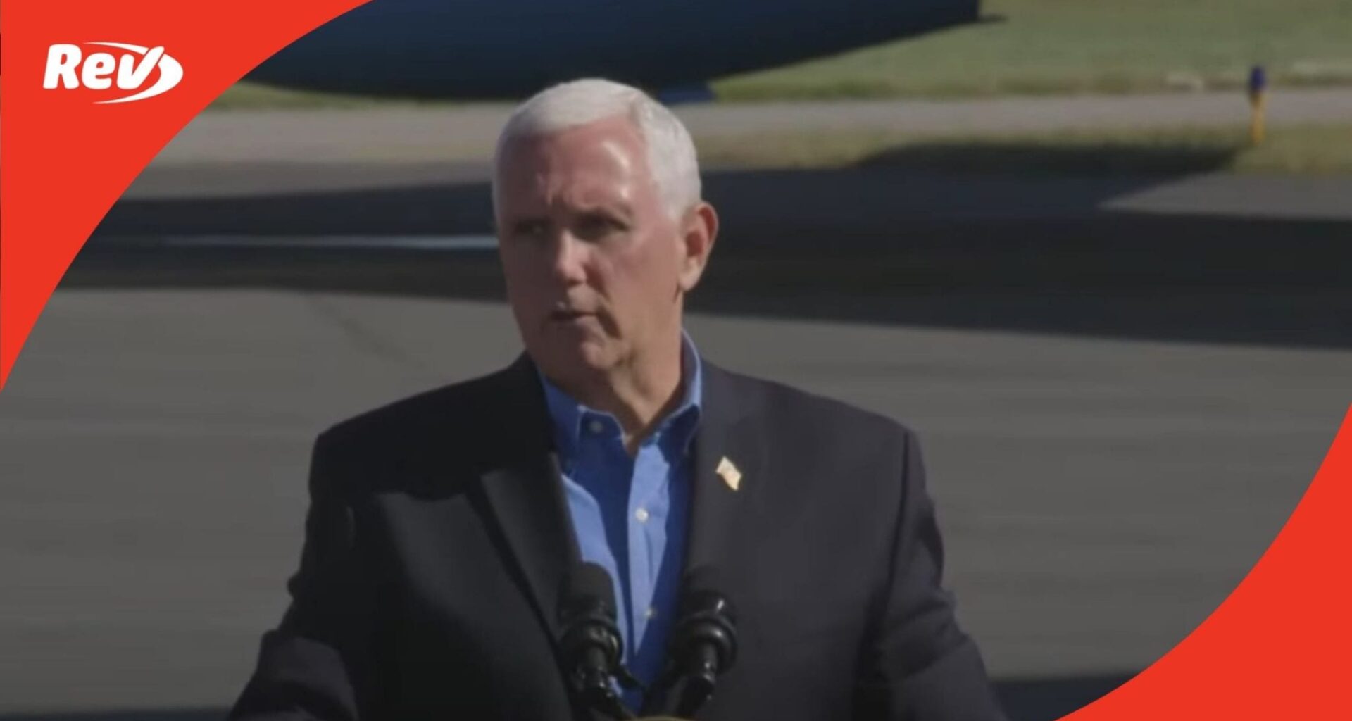 Mike Pence speaking at podium outdoors on airport tarmac, blue shirt, flag lapel pin, Rev logo
