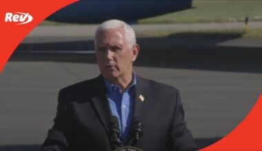 Mike Pence speaking at podium outdoors on airport tarmac, blue shirt, flag lapel pin, Rev logo