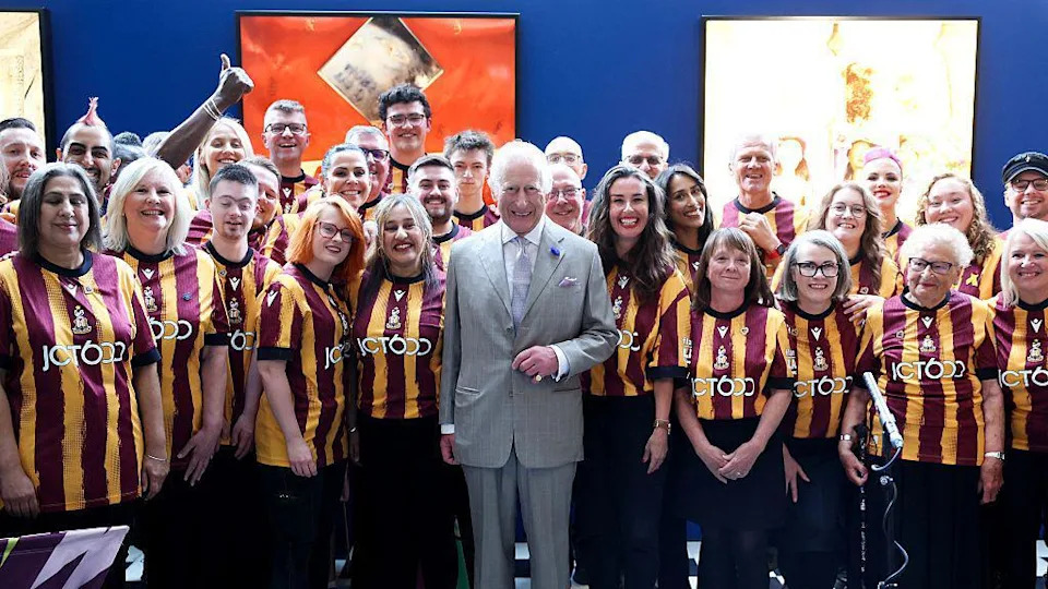 King Charles in the centre of a crowd of people who were members of the Bantam of the Opera choir in Bradford. He is dressed in a grey suit and is smiling; members of the choir are dressed in Bradford City home shirts, which are amber and maroon.