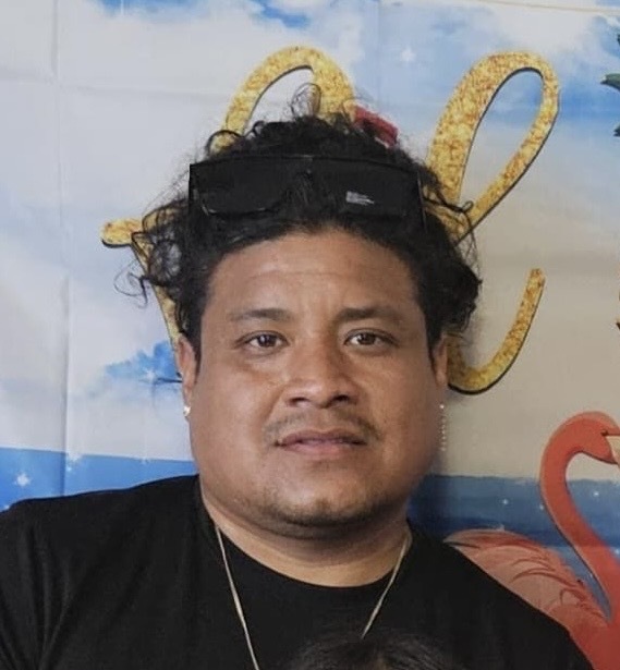 A man with curly hair, sunglasses on his head, and a black shirt stands in front of a decorative backdrop with yellow script and a flamingo.