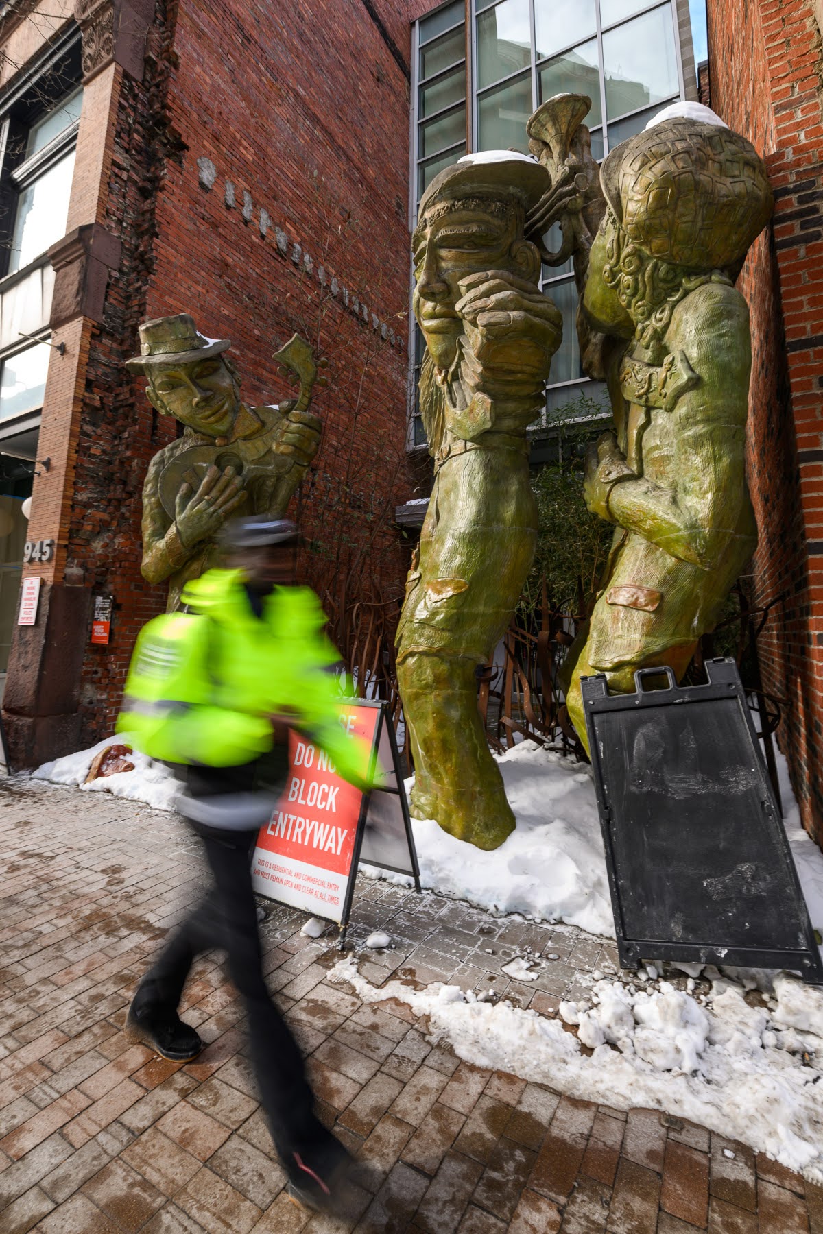 A person in a neon jacket walks past large bronze jazz musician statues outside a brick building on a snowy sidewalk.