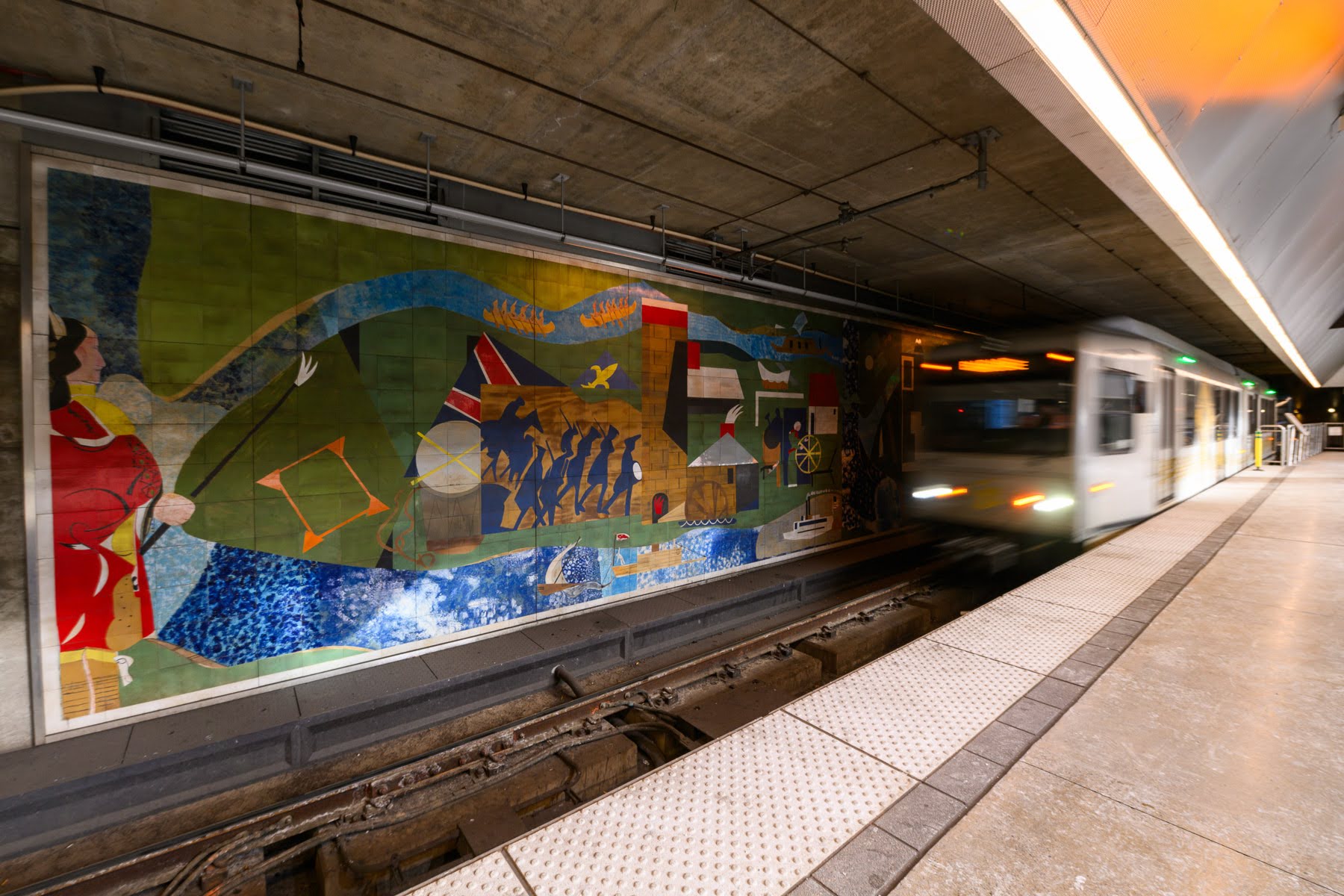 A subway train arrives at an underground station, passing by a large, colorful mosaic mural on the station wall.