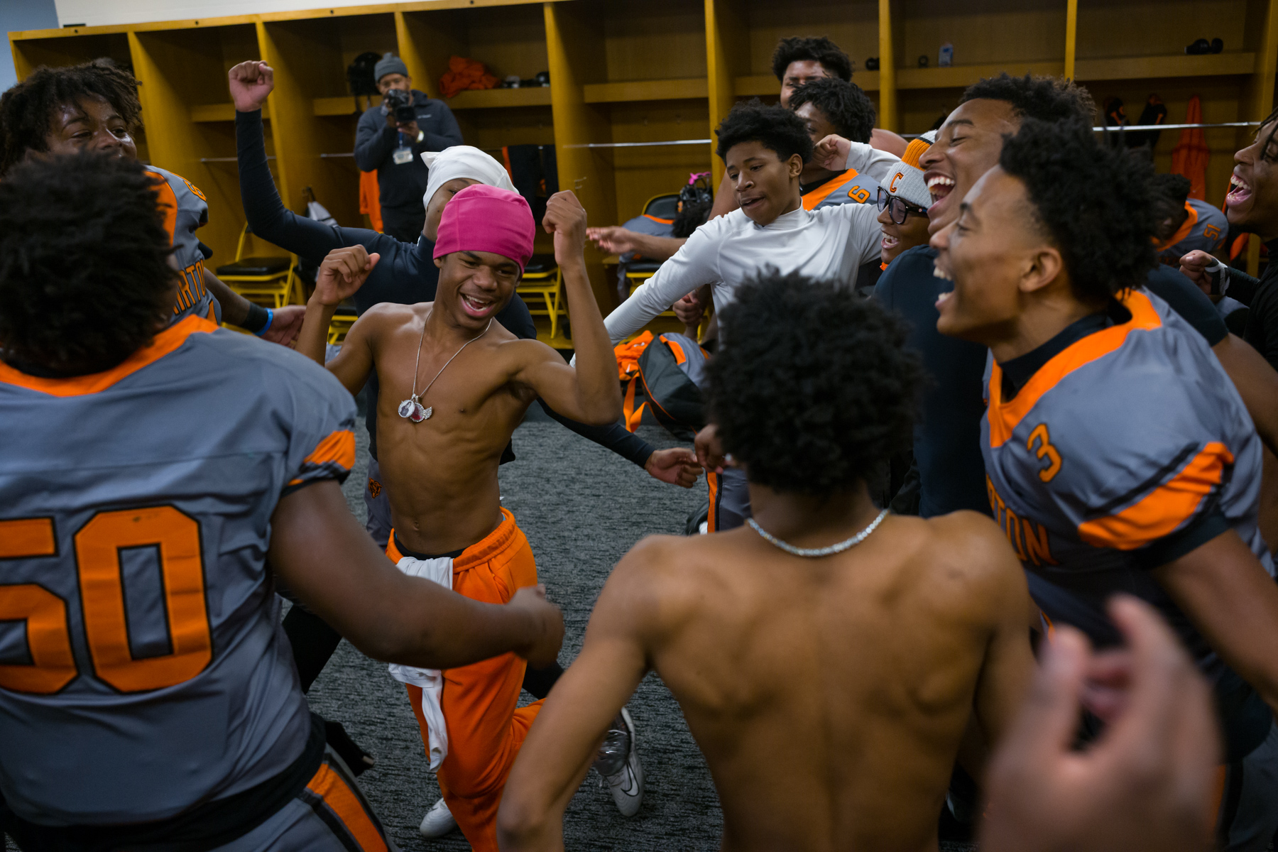 A group of teenage football players in uniforms cheer and celebrate energetically in a locker room.