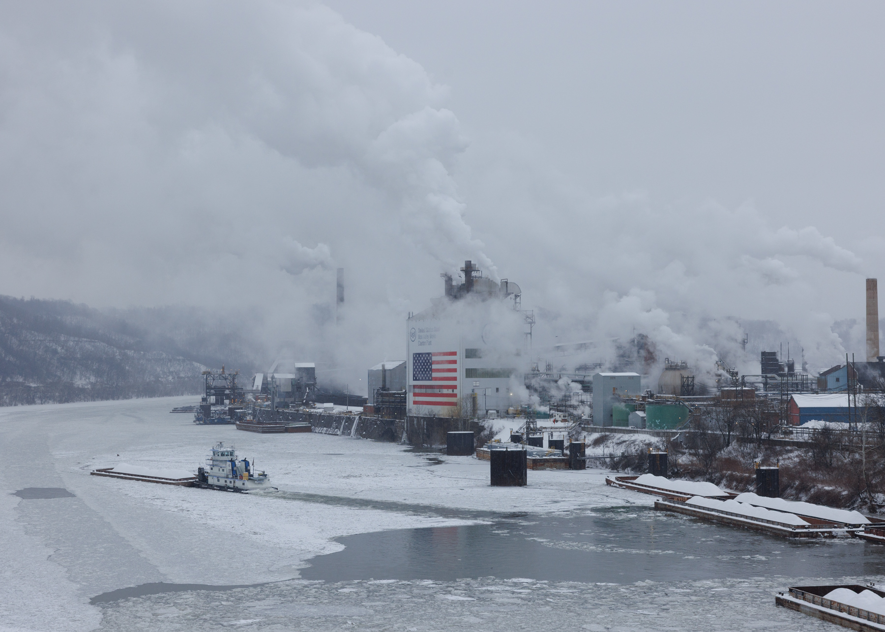 Industrial factories emit smoke near a frozen river, with a large American flag on one building and a boat navigating through the ice. Snow covers the ground and rooftops.
