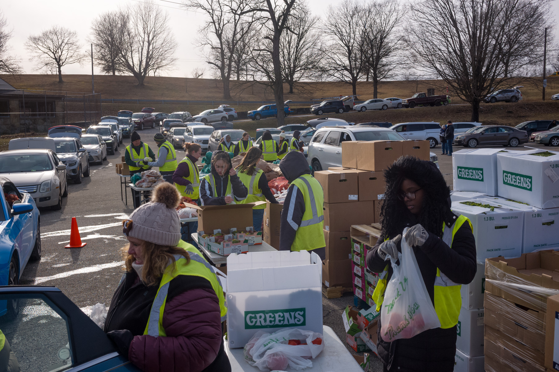 Volunteers wearing yellow vests distribute food and supplies from tables to people in cars at an outdoor drive-through event.