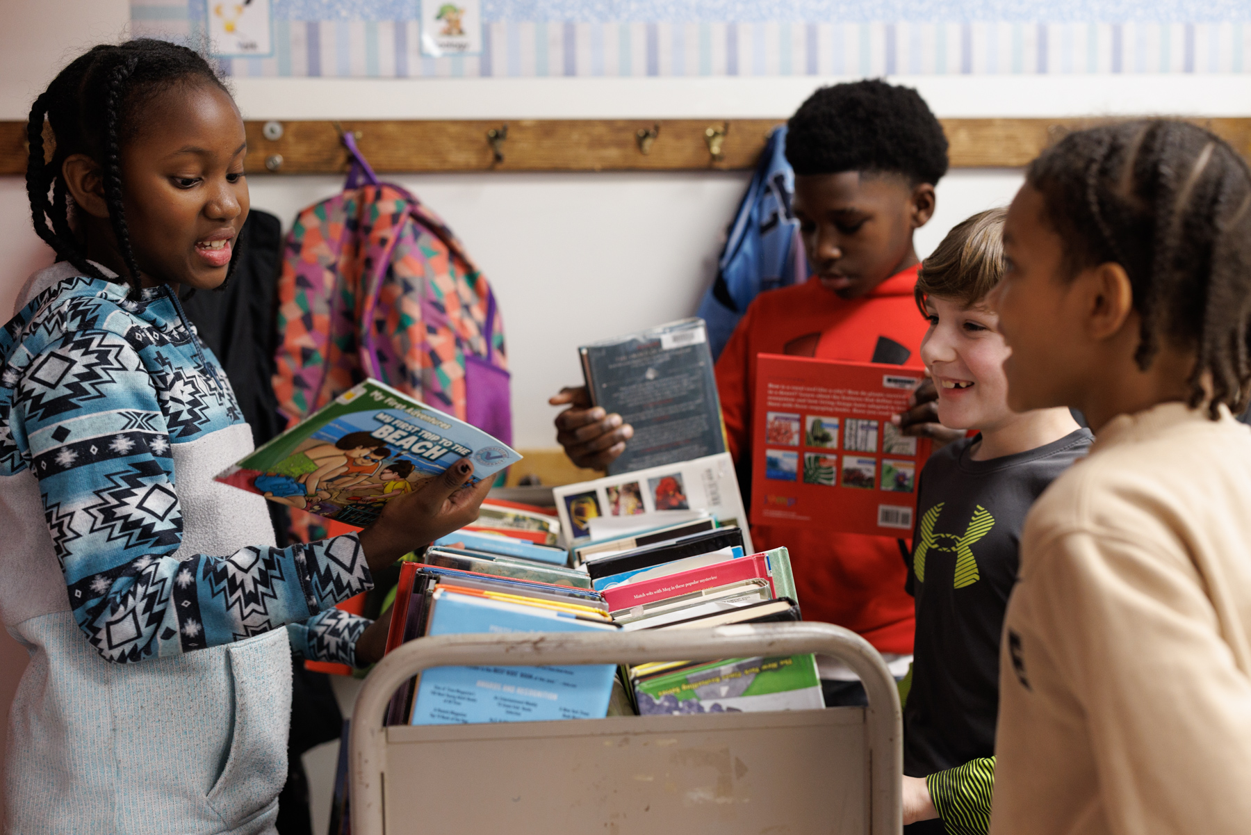 Four children stand around a book cart, selecting and discussing books, with backpacks hanging on hooks in the background.
