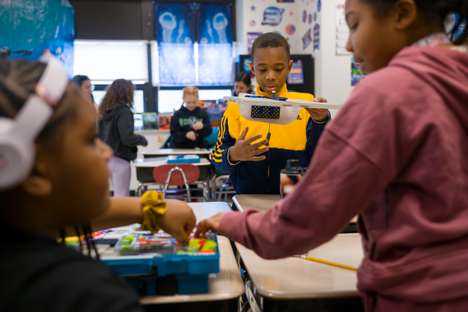 Students work on a hands-on science project in a classroom, with one boy holding a model and others handling materials at their desks.