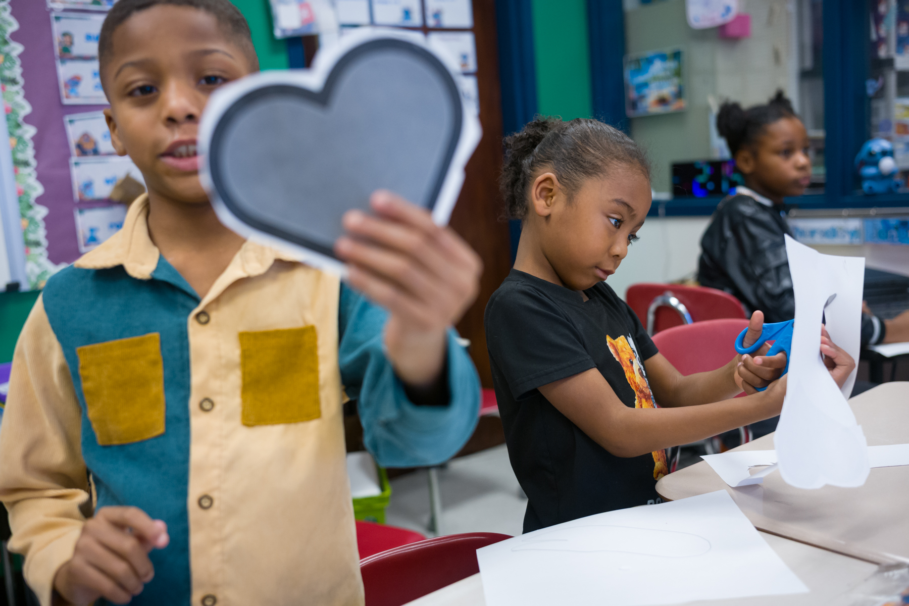 Three children in a classroom work on an arts and crafts project; one holds a cutout heart, another cuts paper, and a third sits in the background.