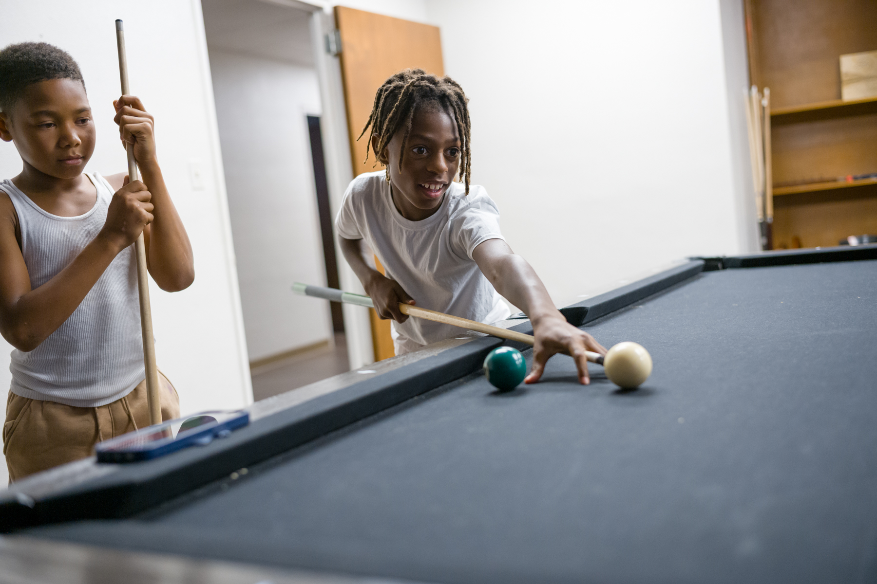 Two boys stand by a pool table; one lines up a shot with a cue stick while the other looks on holding his own stick.
