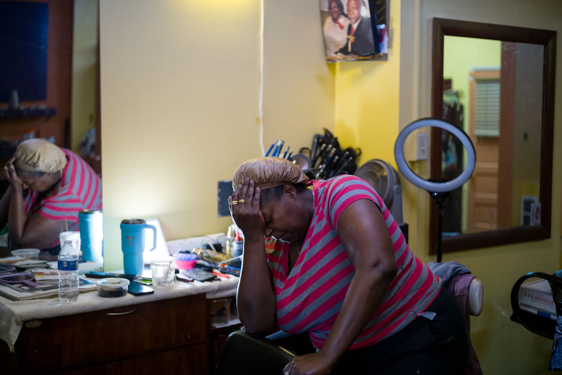A woman in a striped shirt sits in a salon with her head in her hand, appearing distressed. A cluttered counter, mirror, and various salon items are visible in the background.