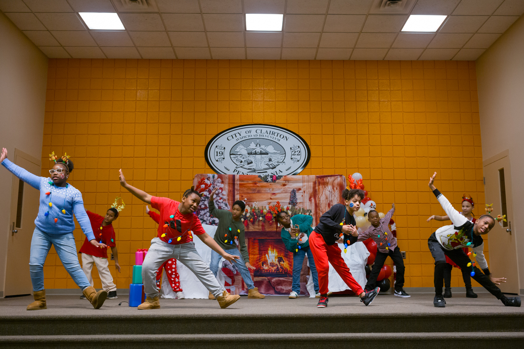 A group of children wearing festive holiday outfits perform a choreographed dance on a stage in front of a winter-themed backdrop.