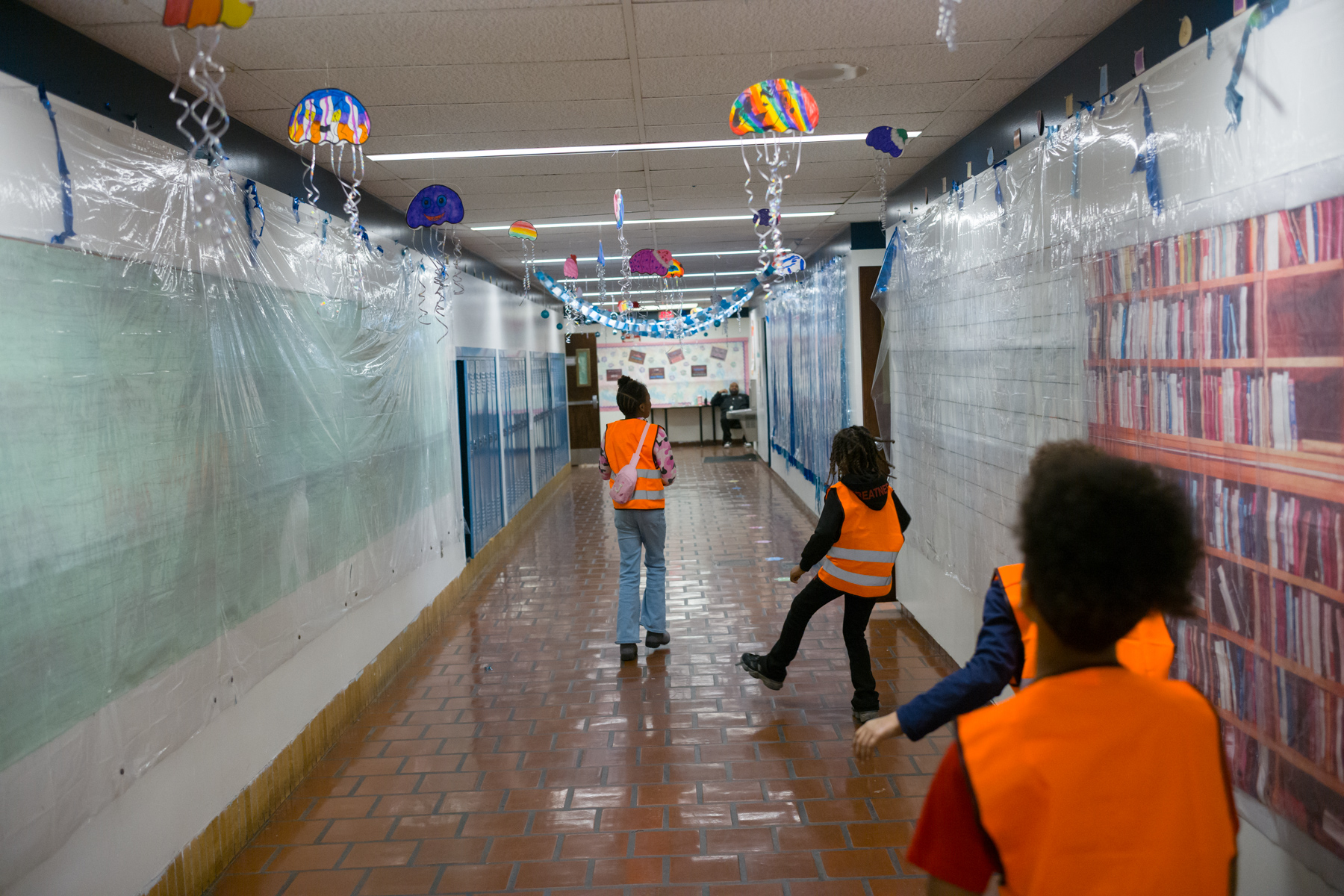 Three children wearing orange safety vests walk down a school hallway decorated with hanging jellyfish and ocean-themed decorations.