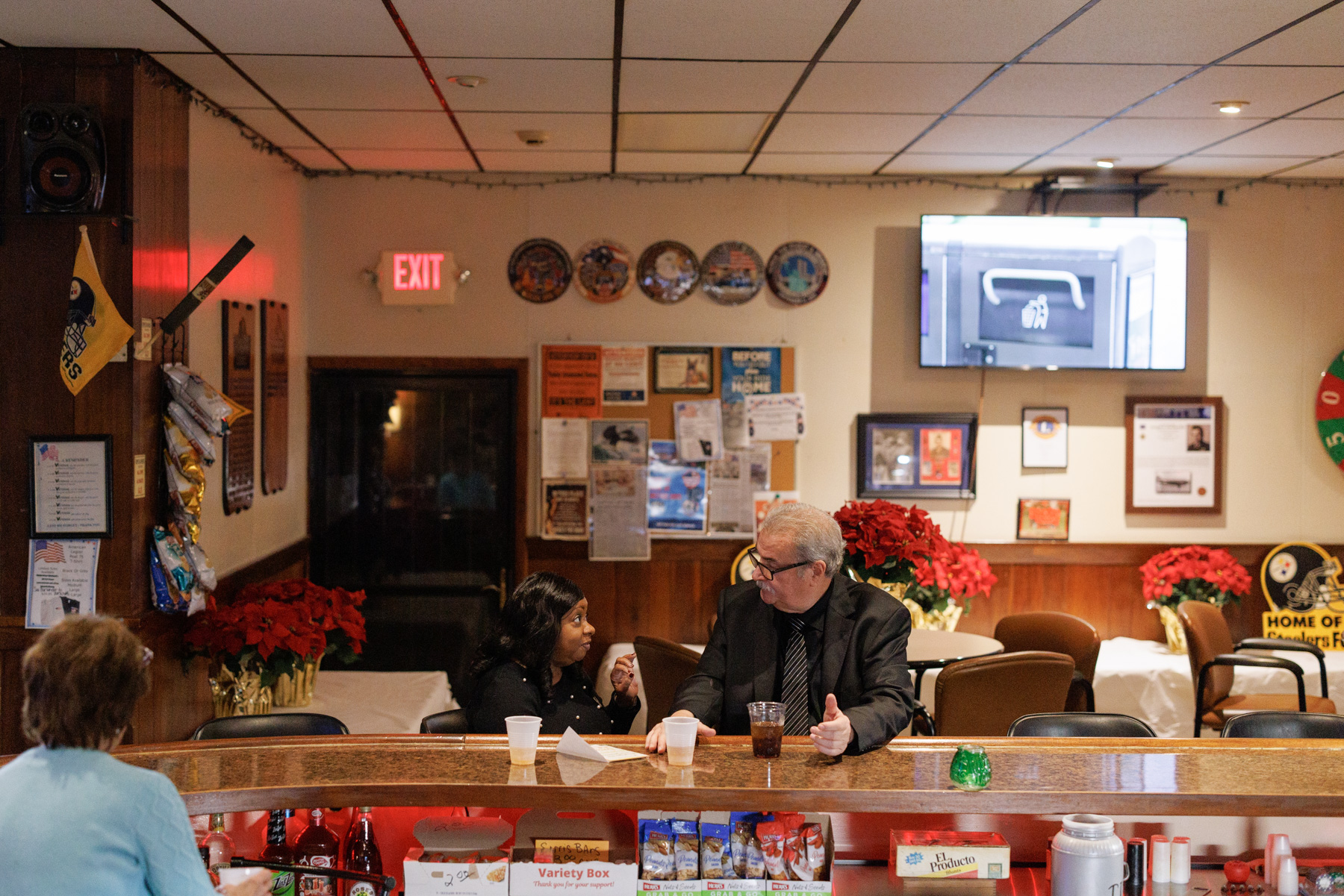 Three people sit at a bar counter talking; bulletin boards, poinsettias, and a TV showing a safe are visible in the background.