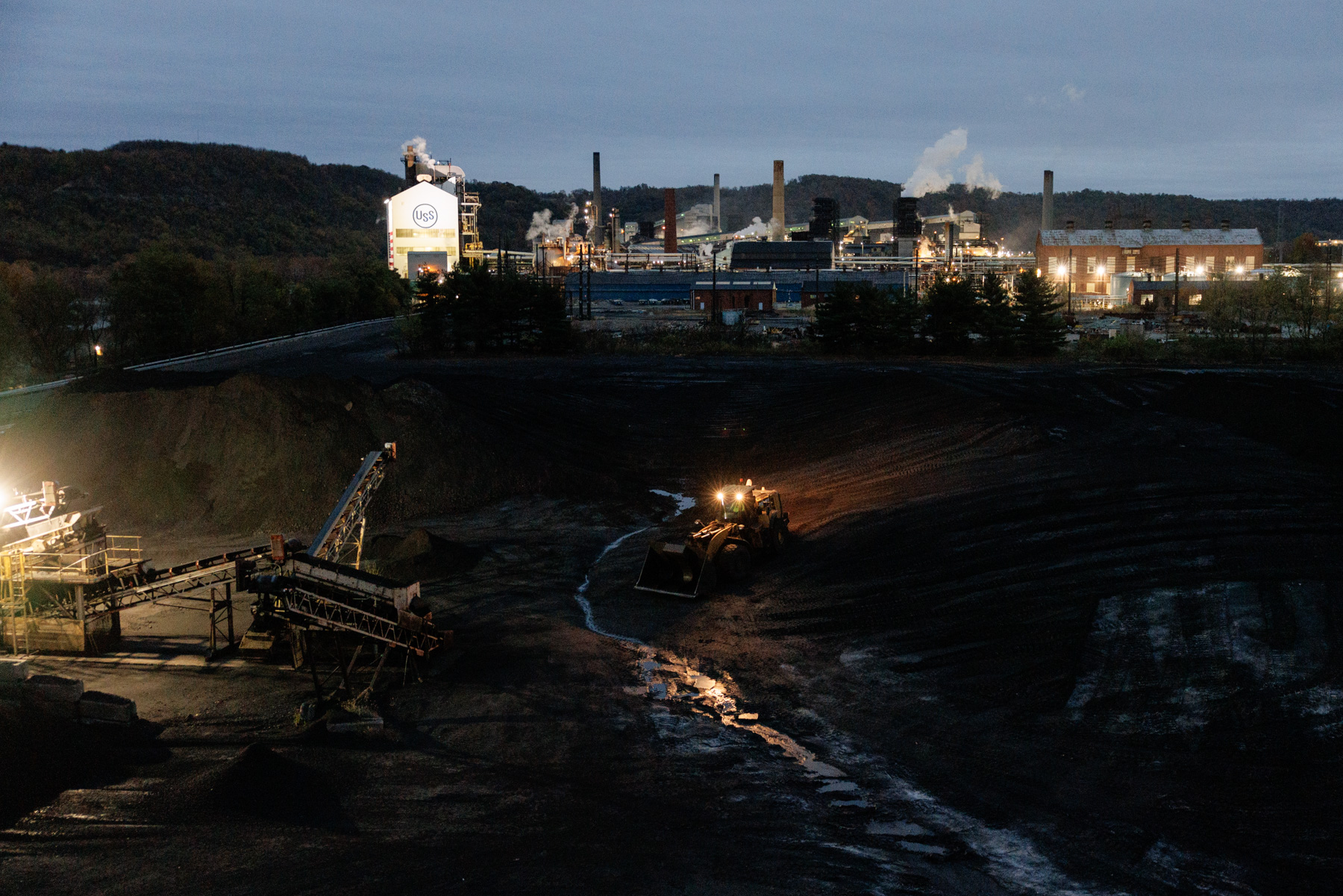 An industrial plant emits smoke in the background while heavy machinery operates on a dark, excavated landscape in the foreground under cloudy sky.