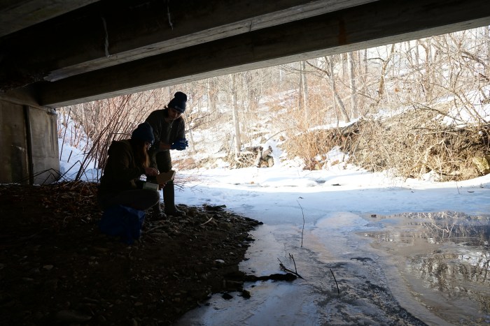 Two people stand under an overpass along a stream.