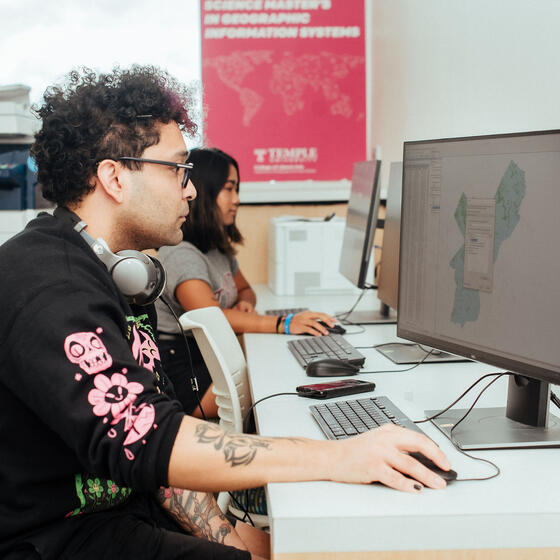 Two people sit at desks using desktop computers, with maps displayed on the monitors. A Temple University poster is visible on the wall in the background.