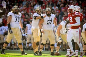 College football players preparing for a snap during a game, wearing white and red uniforms on a sunny field.