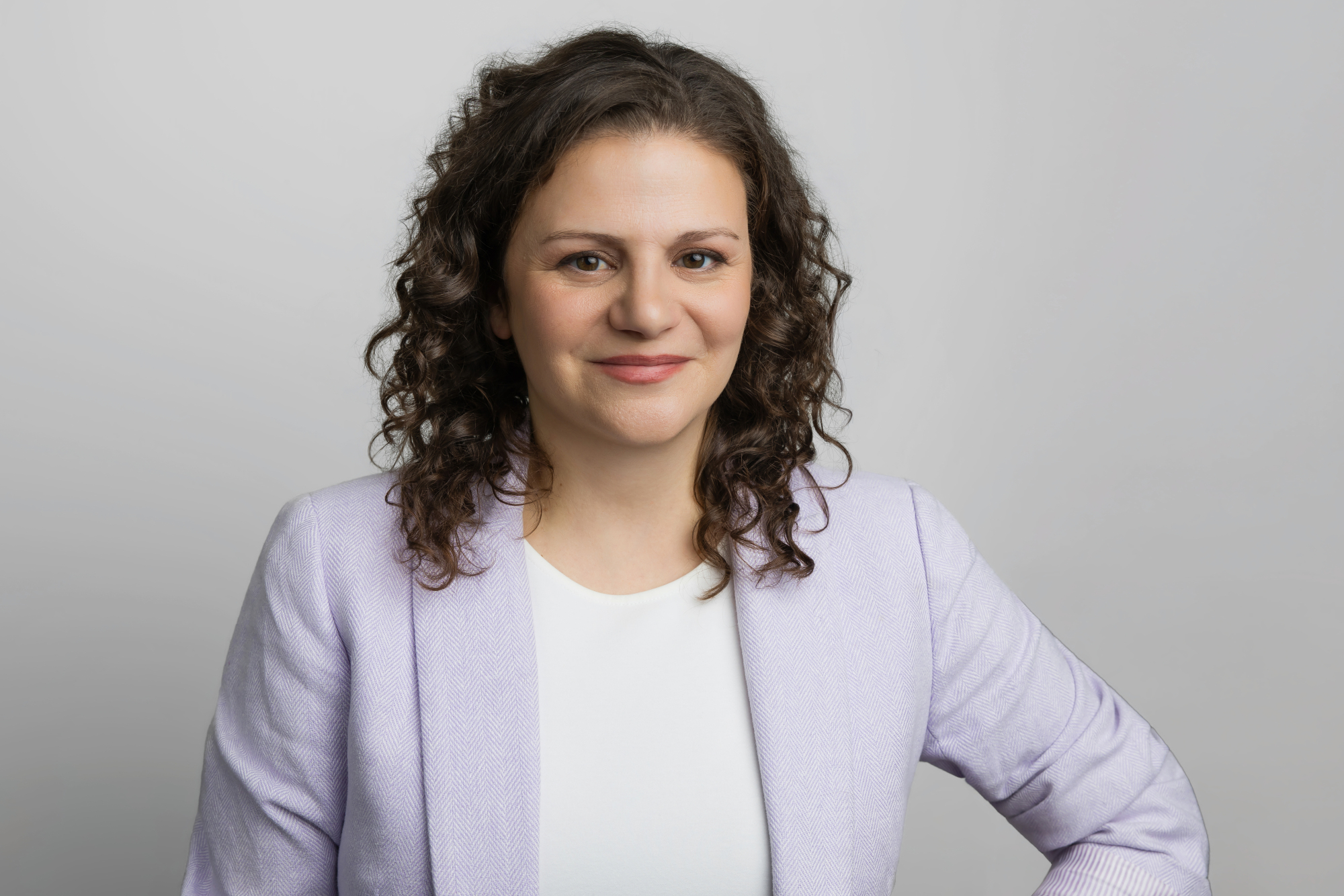 Person with curly brown hair wearing a light purple blazer and white top, smiling, posed against a plain light gray background.