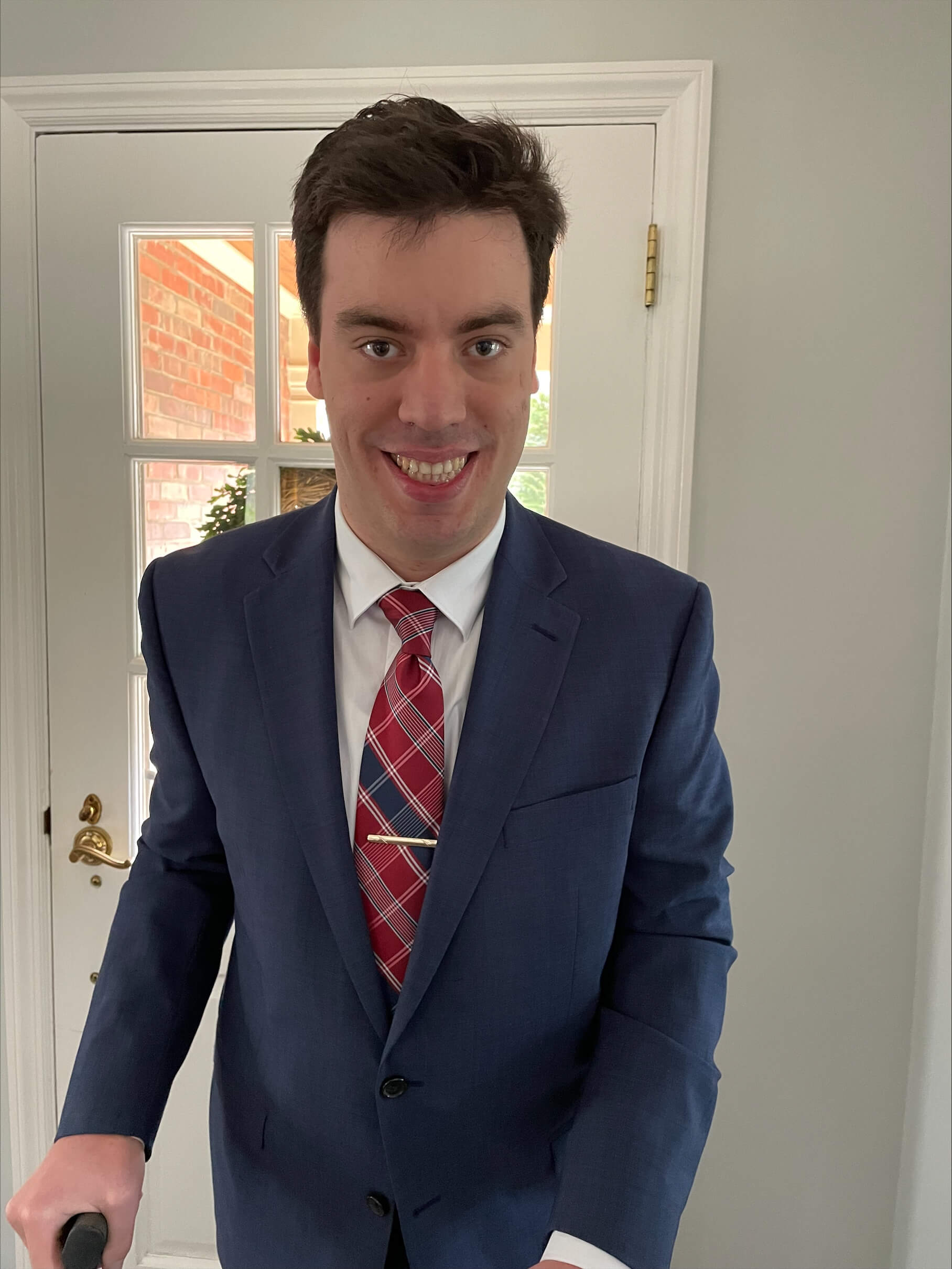 A person in a blue suit, white shirt, and red plaid tie stands indoors in front of a white door, smiling at the camera.