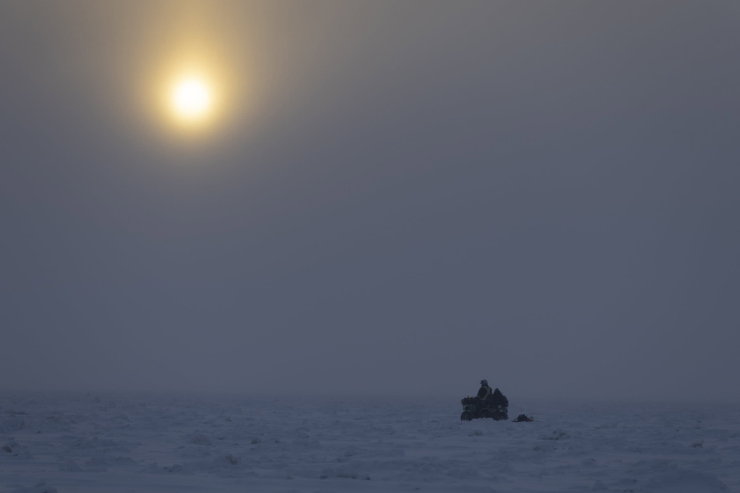 A person on an ATV on a frozen lake under a morning sun in fog