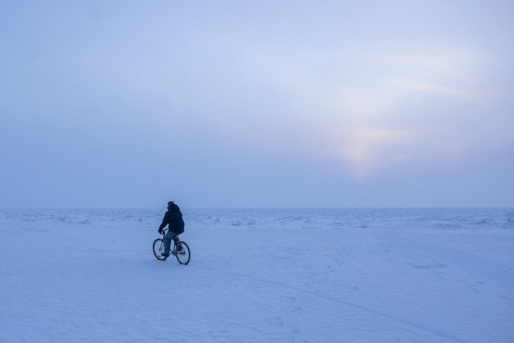 A person rides a bike on a frozen lake under morning sunrise with blue hues