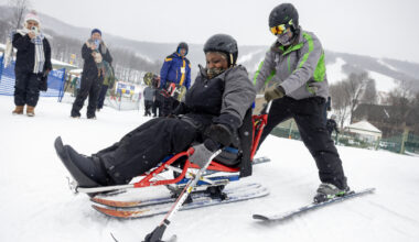 A woman is seated on an adaptive sled, while a ski instructor in winter clothing and on skis holds onto the back of the sled. They are on snow, and a few people look on from the background.