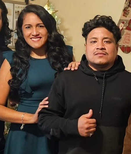 A woman in a teal dress stands next to a man in a black hoodie giving a thumbs up, both posing indoors and smiling at the camera.