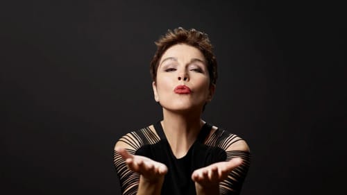 Andreas, a white woman with short brown hair, dramatically blows a kiss at the viewer.