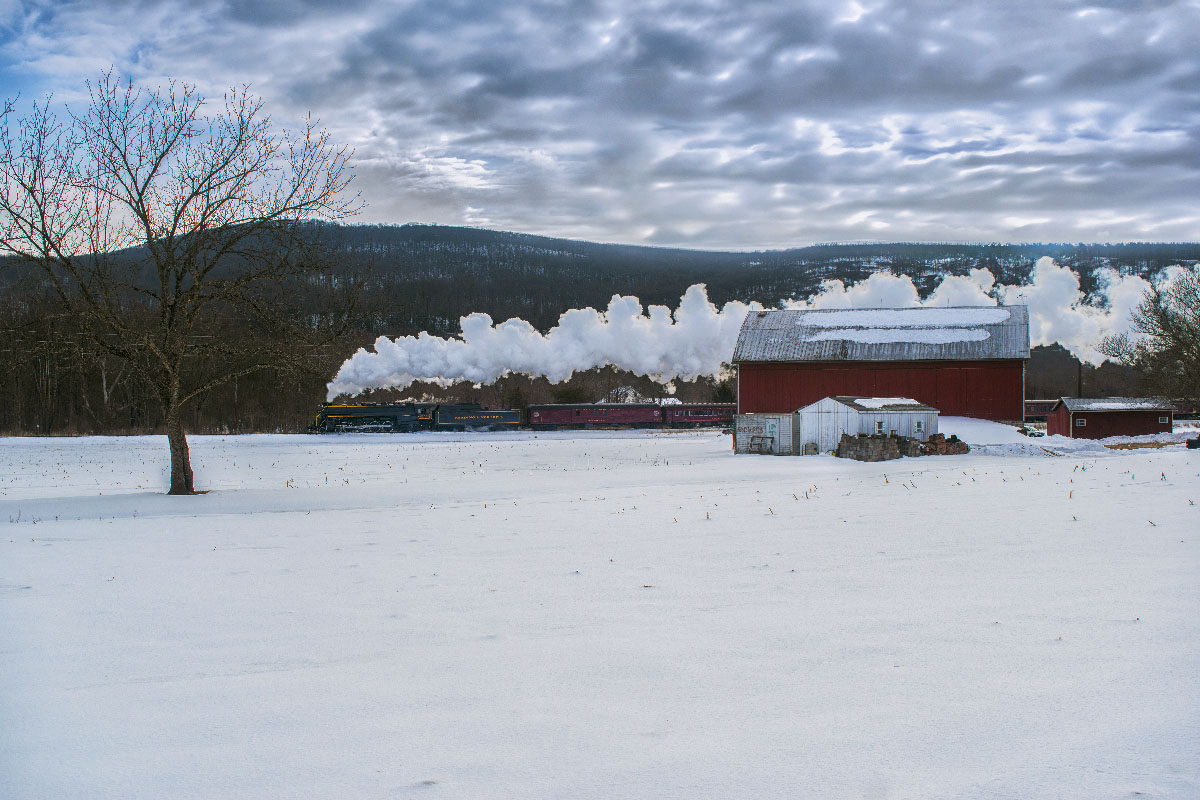 Black steam locomotive passing a red barn in a snowy landscape. Reading & Northern No. 2102 winter rambles.