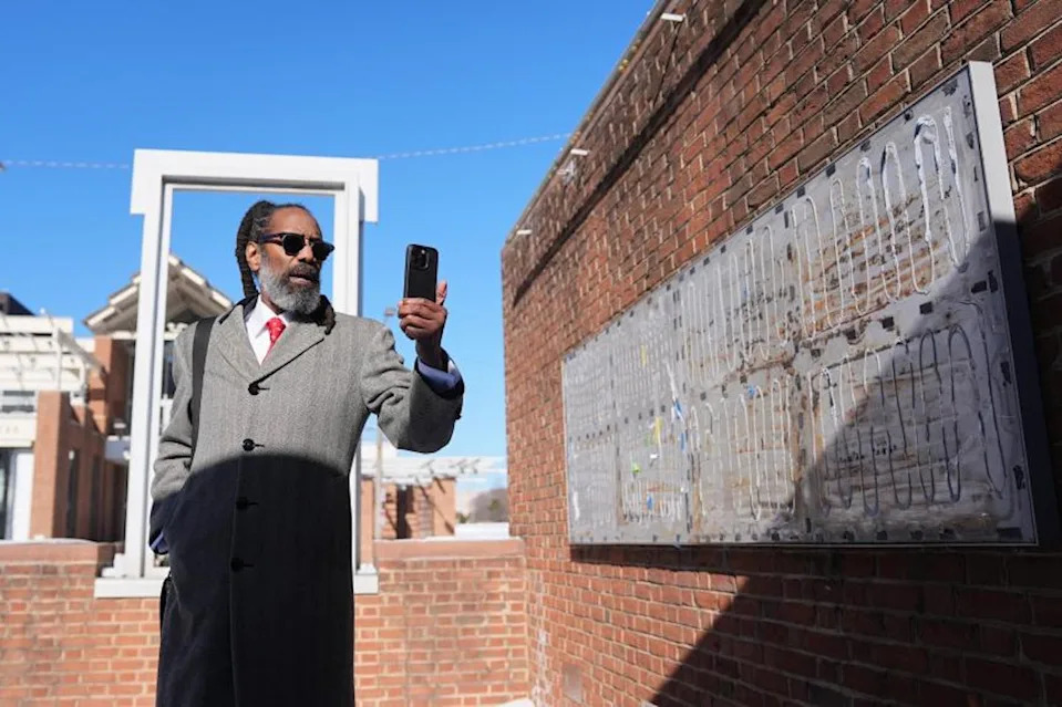 Man in overcoat and sunglasses holds up phone, with brick walls around him