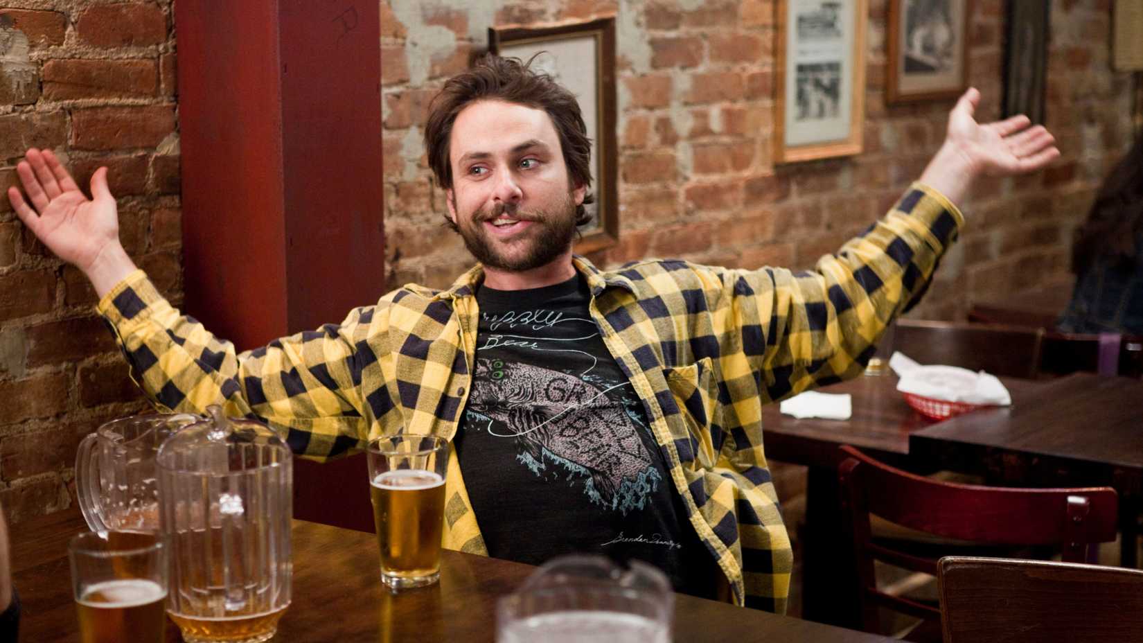 Charlie Day sitting in Paddy's Pub with his hands outstretched and a beer on the table in It's Always Sunny in Philadelphia