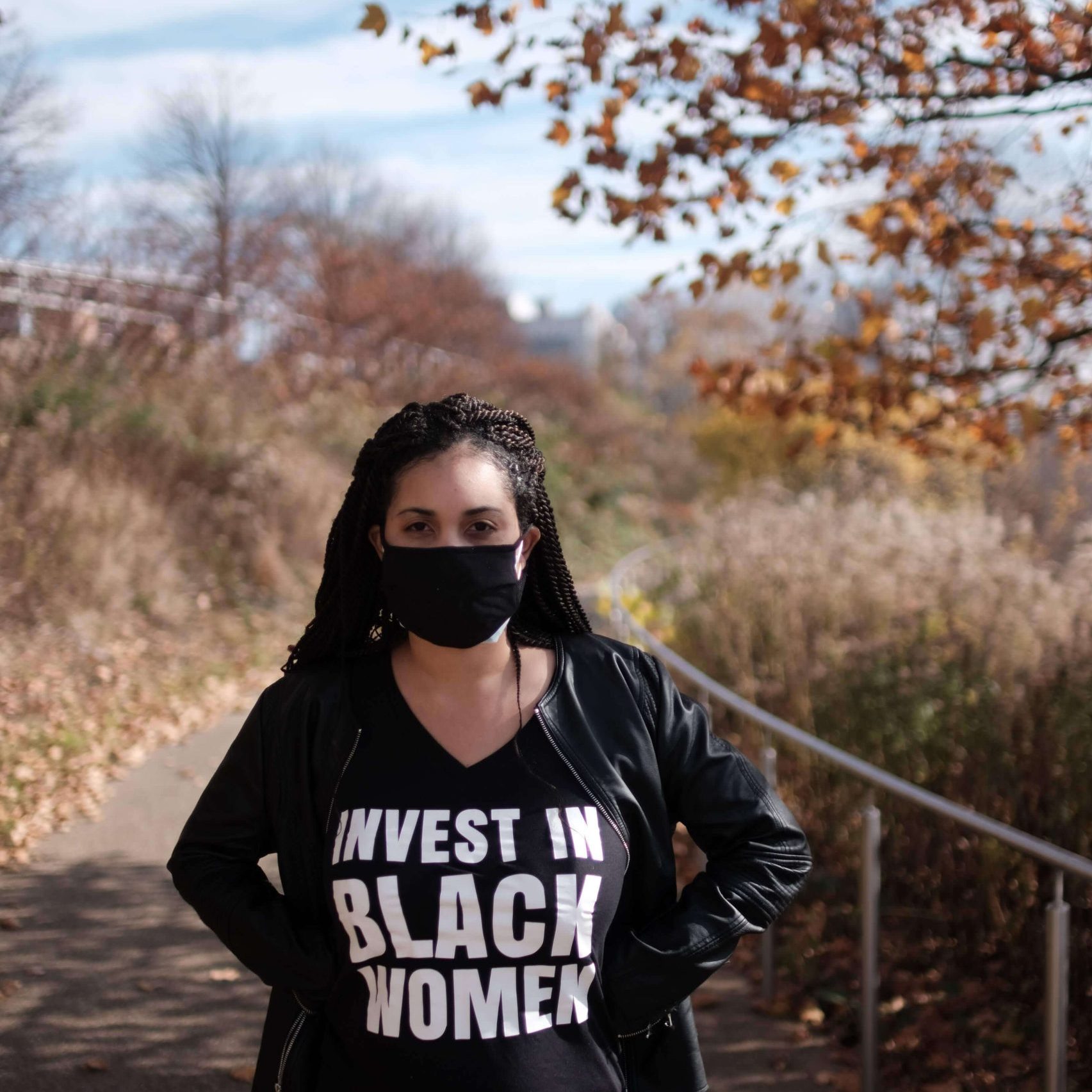 A person stands outdoors on a path, wearing a black mask and a T-shirt that says "Invest in Black Women," with autumn trees in the background.