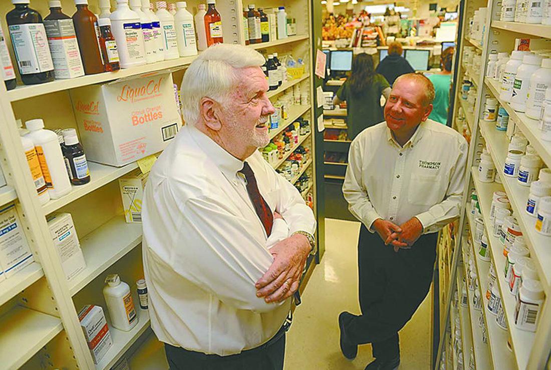 Two men smile while standing next to well-stocked pharmacy shelves