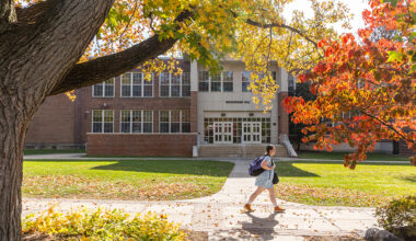 A student walks across a sunlit campus walkway in front of a brick academic building, surrounded by vibrant fall foliage in shades of yellow, orange, and red.