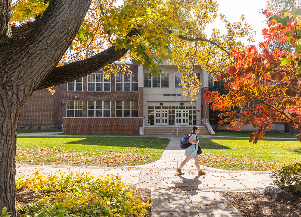 A student walks across a sunlit campus walkway in front of a brick academic building, surrounded by vibrant fall foliage in shades of yellow, orange, and red.
