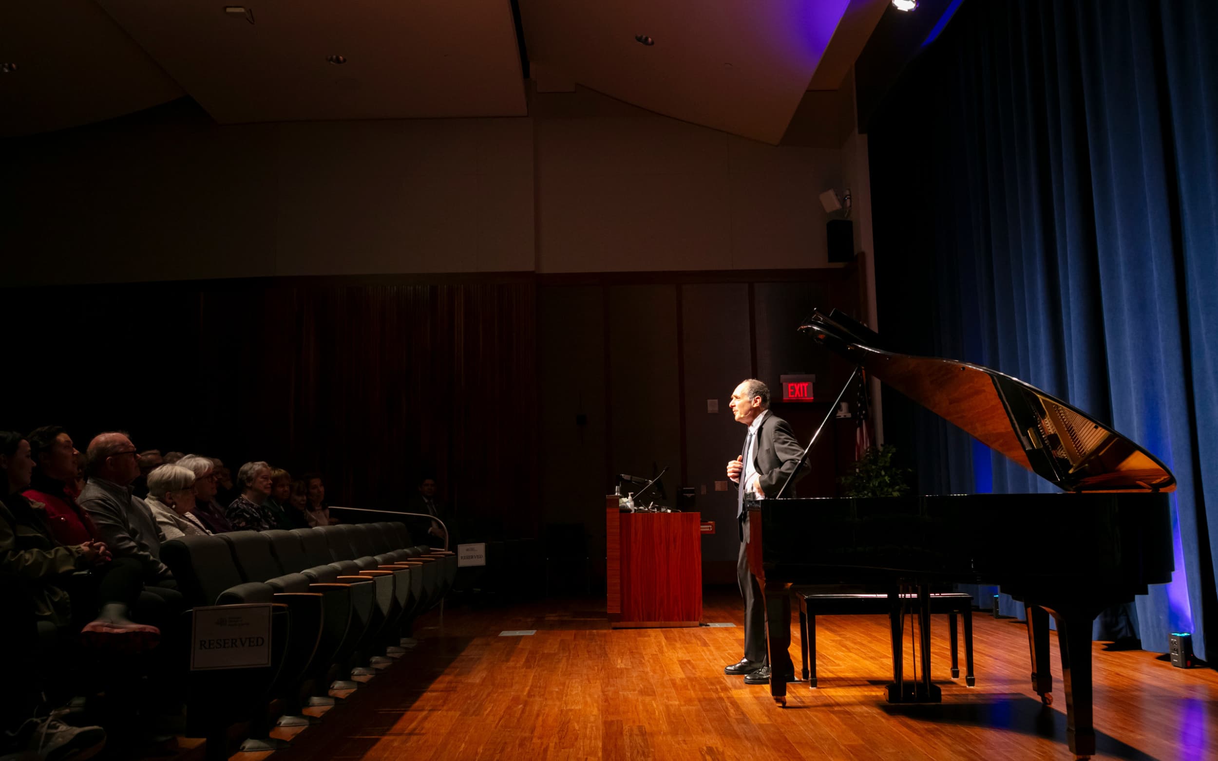 Speaker on stage beside a grand piano, addressing an audience in an auditorium.