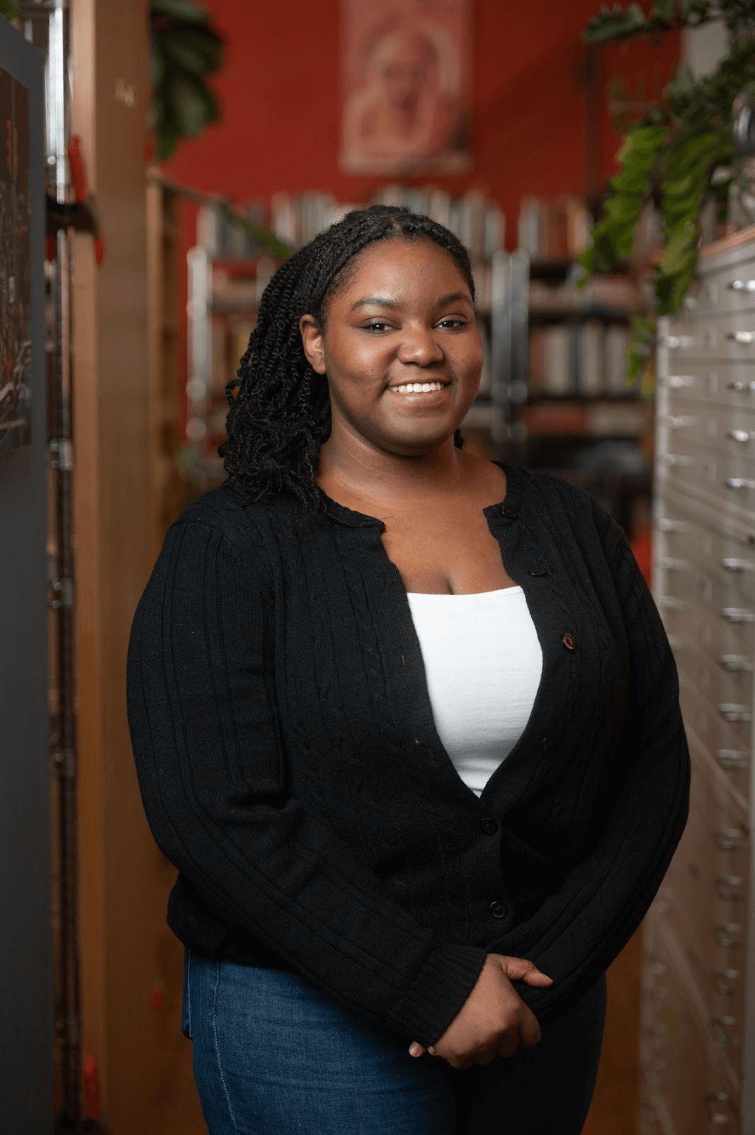 A woman with braided hair, wearing a black cardigan and white top, stands indoors in front of bookshelves and drawers, smiling at the camera.