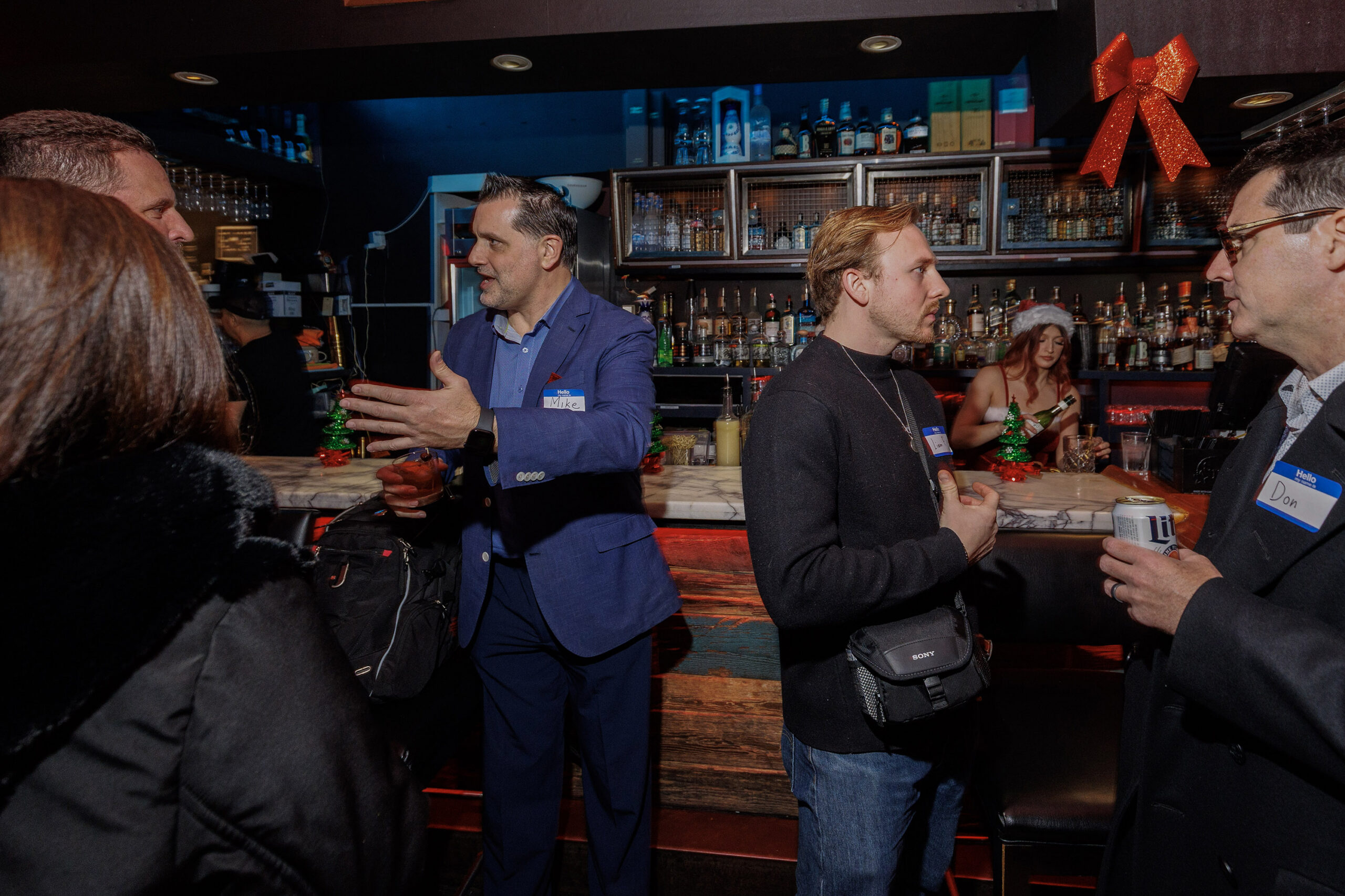 Several people talk and mingle at a bar decorated with holiday ornaments; a bartender in a Santa hat prepares drinks in the background.