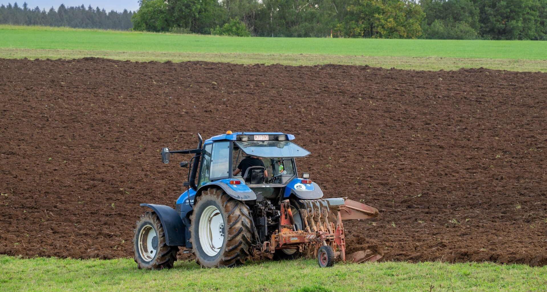 A blue tractor plowing a large, freshly-tilled field with green trees in the background.