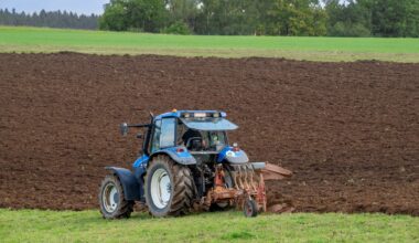 A blue tractor plowing a large, freshly-tilled field with green trees in the background.