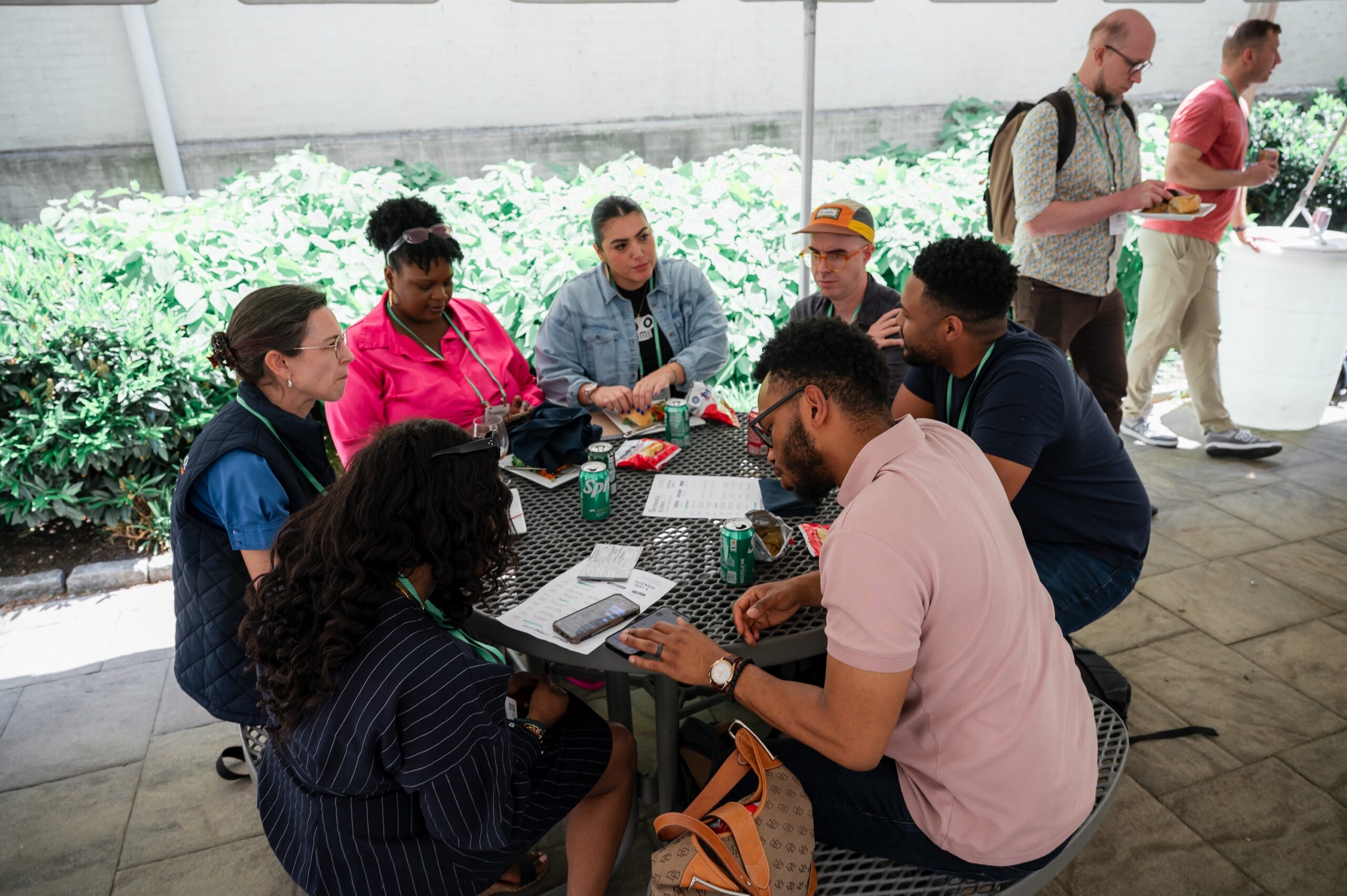 A group of seven people sits around an outdoor table having a discussion, with papers and drinks on the table; two other people stand nearby.