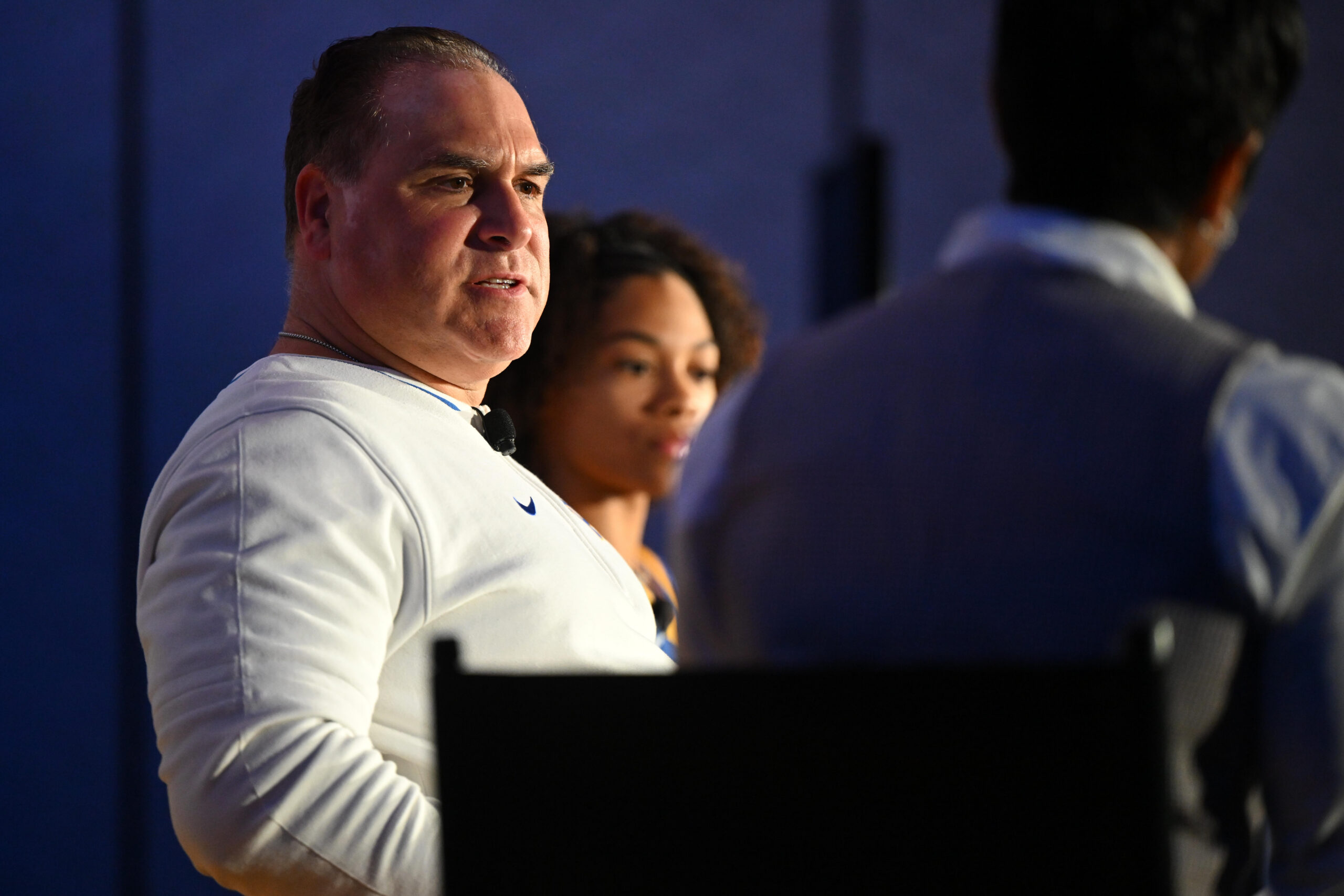 Pittsburgh head coach Tory Verdi sits in a chair on a stage with one of his players in the background and an interviewer in the foreground. He's wearing a white long-sleeve shirt.