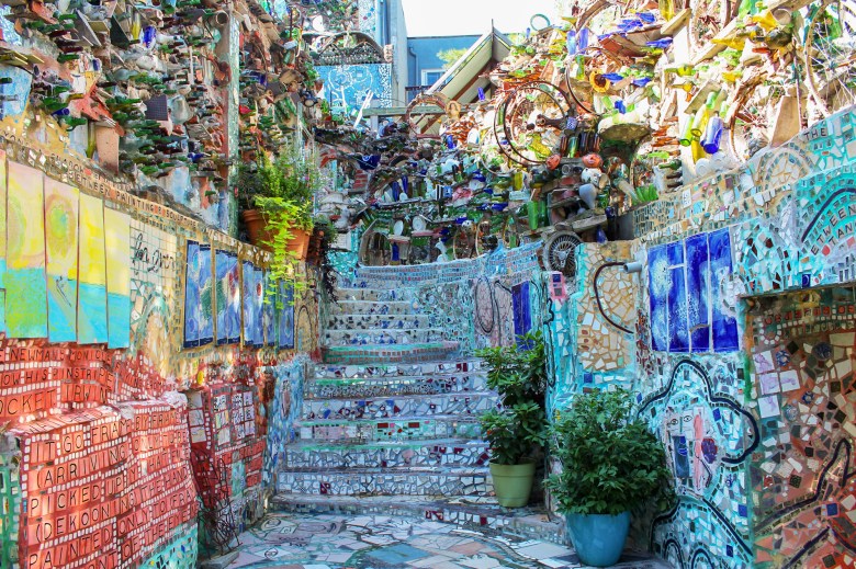 A view of a large staircase outside at Philadelphia's Magic Gardens. The mosaicked staircase leads up and is flanked by flat tiled walls. At the top of the stairs the walls continue and become more sculptural, including bicycle wheels, bottles, and ceramic works stacked together.