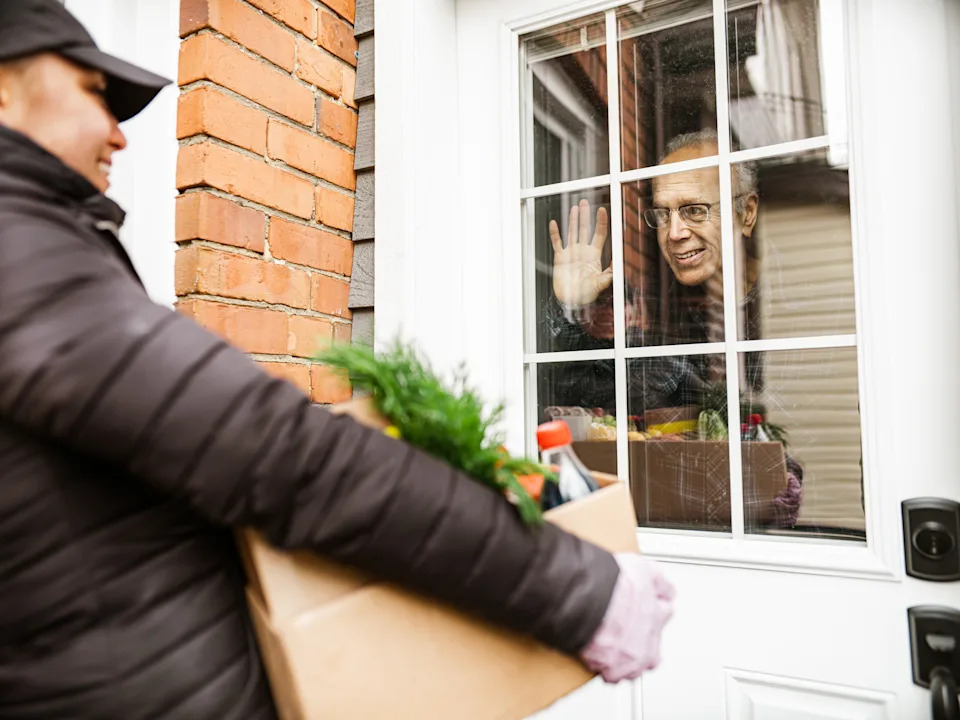 Person inside waves while someone delivers groceries outside a window