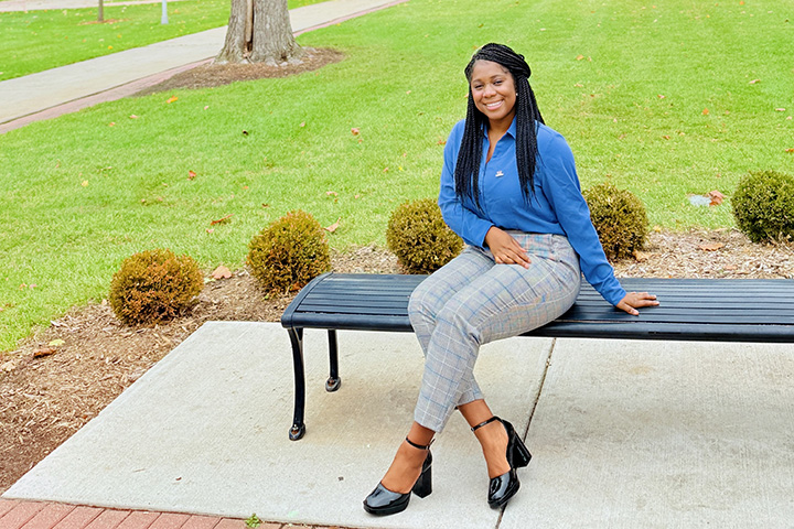 A woman with long black hair and wearing a blue blouse and checker-pattern pants sits on a blue-green bench surrounding by grass.