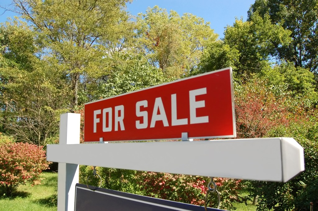A red "FOR SALE" sign with white text, positioned on a white post against a backdrop of green trees and blue sky.