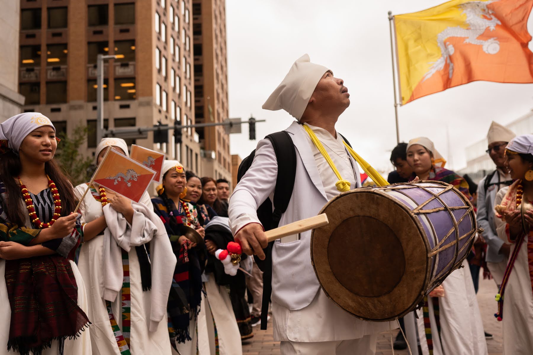 A group of people in traditional attire participate in a street parade; one person plays a large drum while others stand nearby, with a flag visible in the background.