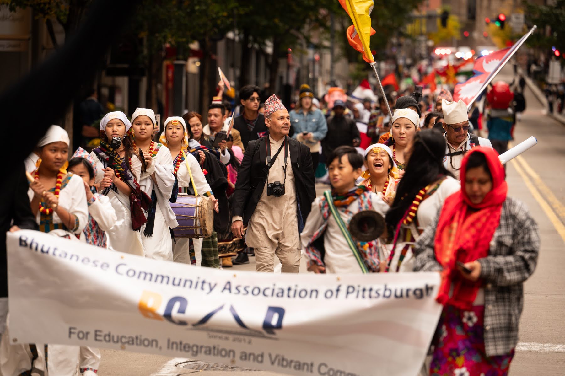 A group of people in traditional attire march down a city street holding a banner that reads "Bhutanese Community Association of Pittsburgh.