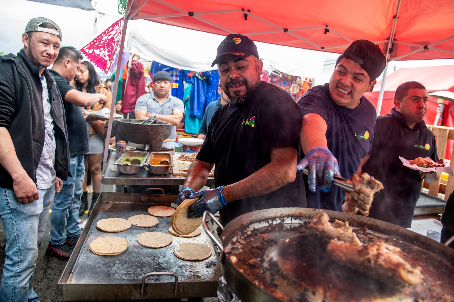 Two men cook meat and tortillas at an outdoor food stall under a red canopy, while others wait in line and watch them prepare the food.