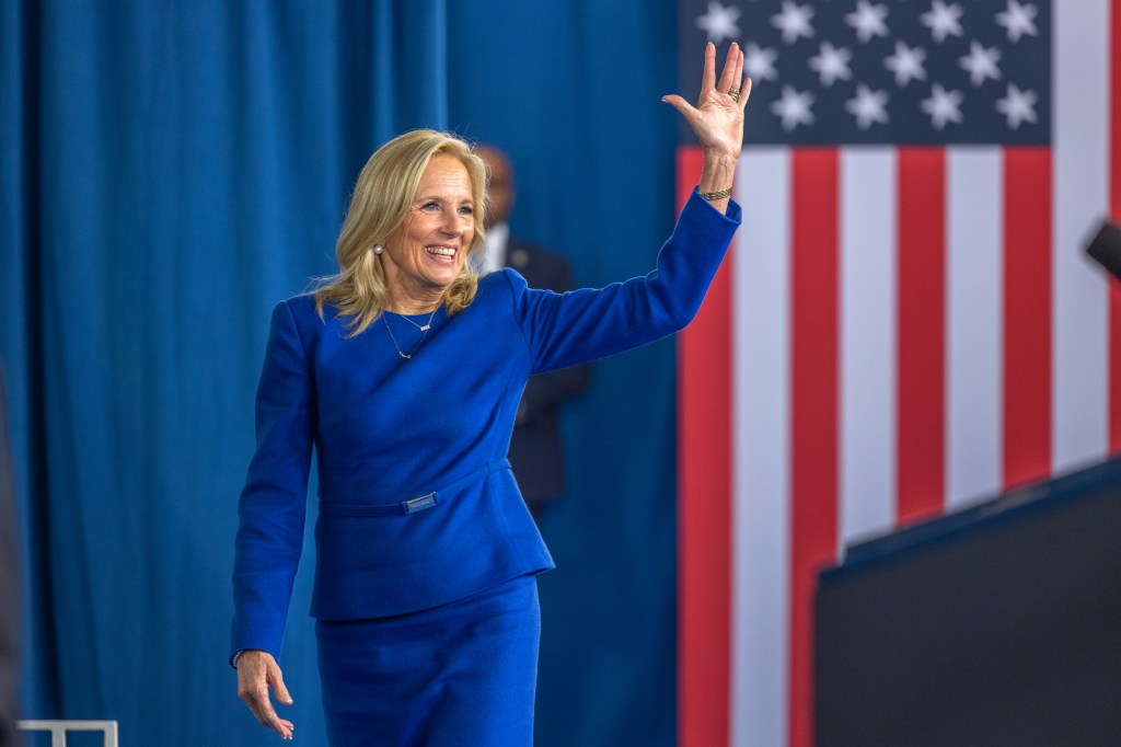 Jill Biden waves to the crowd in a blue suit, in front of a blue backdrop and an American flag.